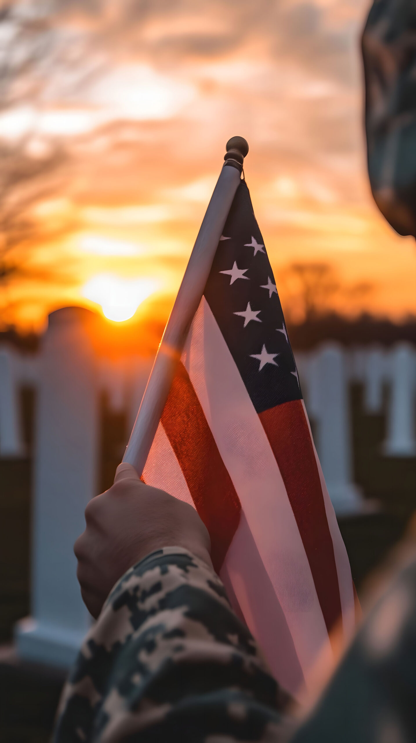 Image of part of a soldier holding up a small American Flag toward a military cemetery with the sunset in the background